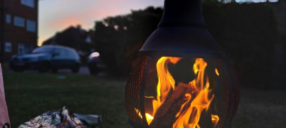 Metal chiminea with a log fire glowing inside, on a garden lawn at dusk