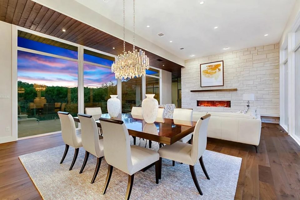 Dining room with a crystal chandelier over a glossy table, white upholstered chairs and a stone fireplace wall