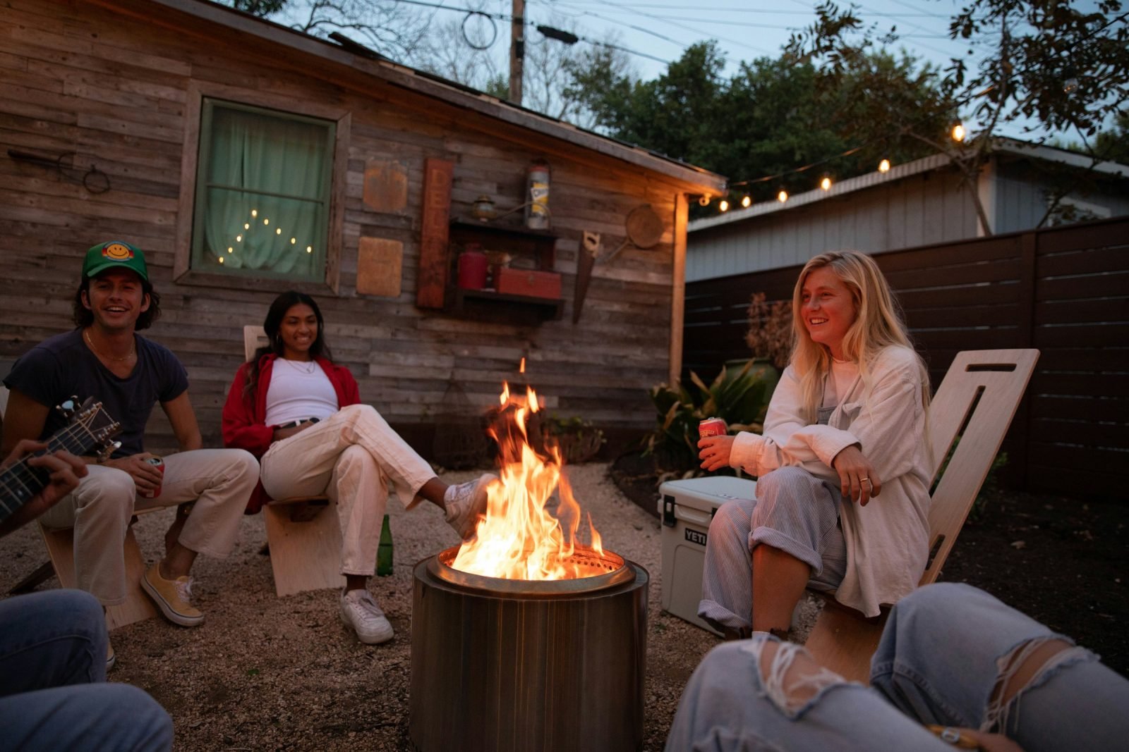 Friends relaxing around a garden fire under string lights in the evening