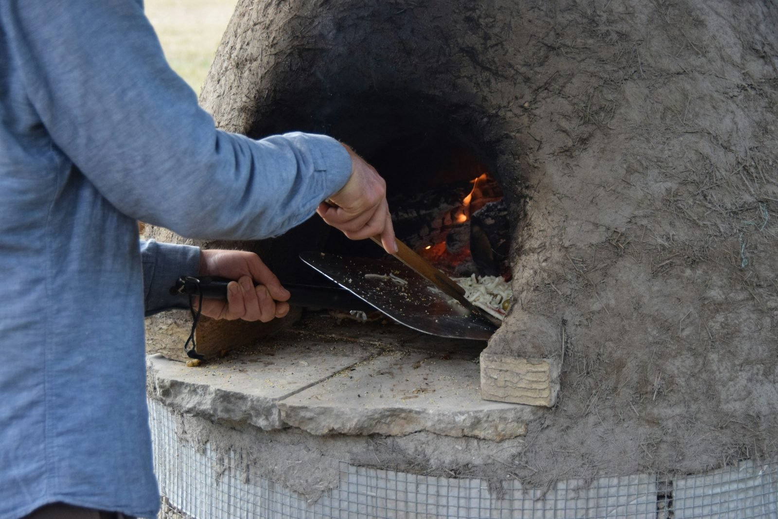 Person sliding a pizza into a clay oven with a metal peel