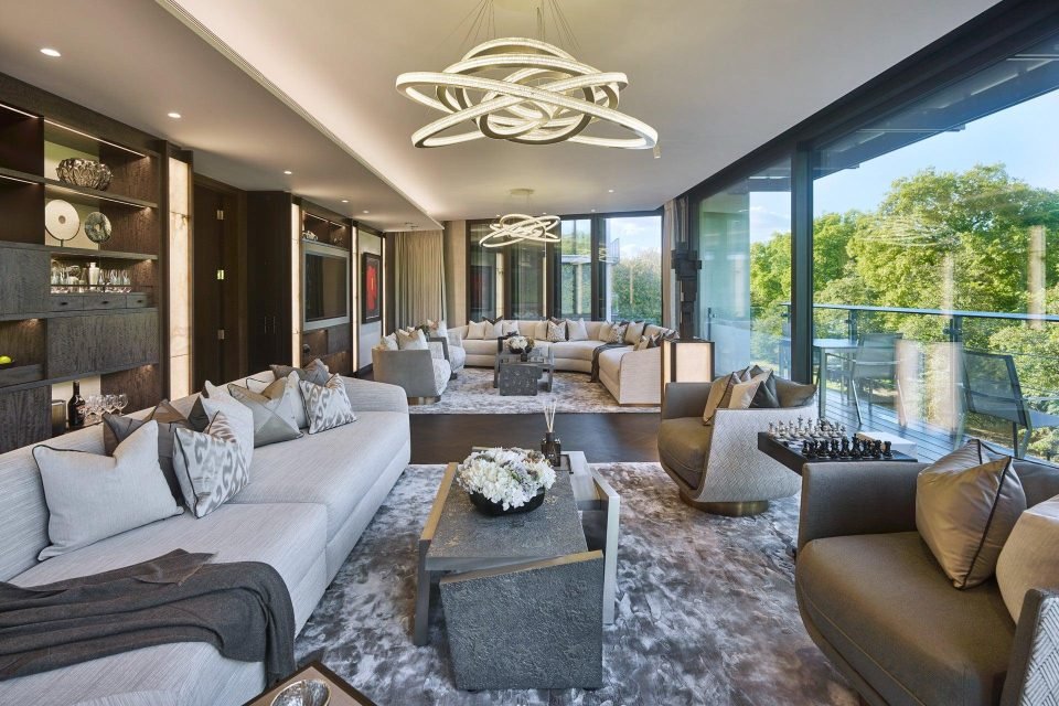 Long open living room with a sculptural ring chandelier, grey sofas, shaggy rug and a glass balcony