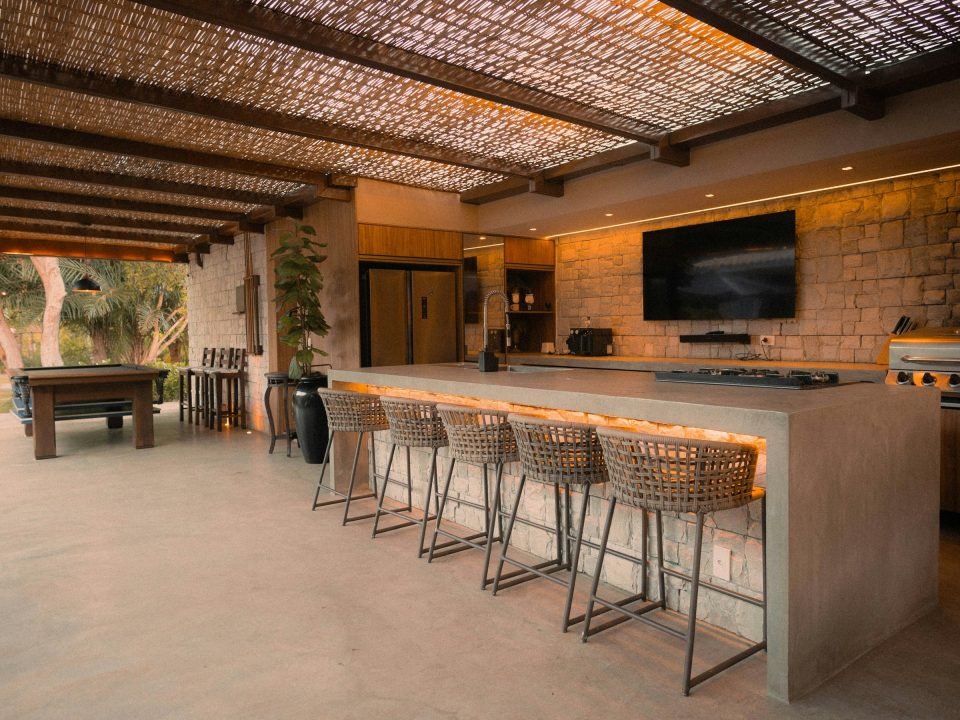 Covered outdoor kitchen with a long concrete island, woven bar stools, a gas hob and a stone feature wall