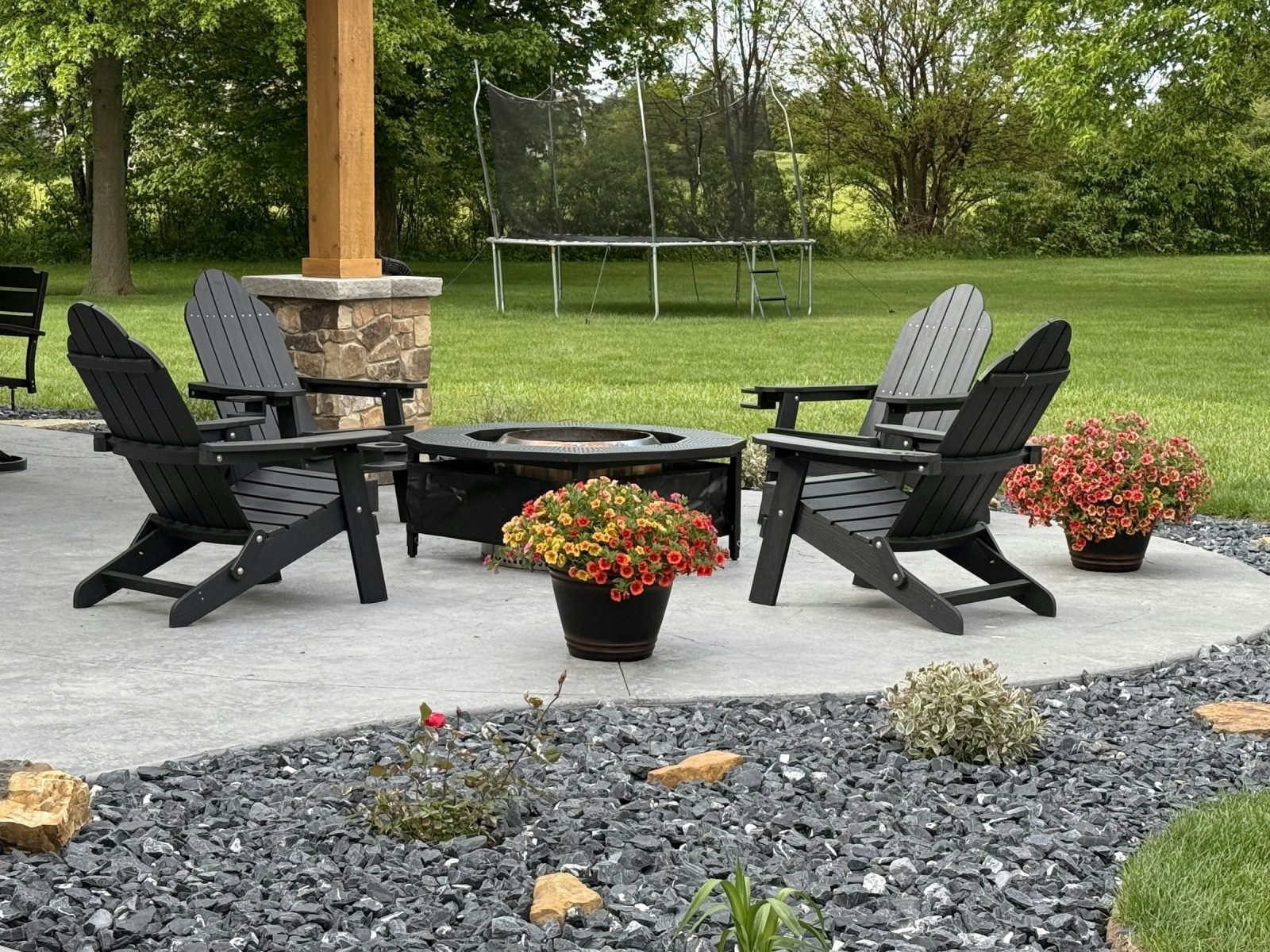 Round fire pit on a concrete patio surrounded by four Adirondack chairs and flowers